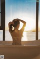 A woman in a white bikini sitting in a bathtub looking out a window.