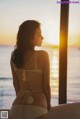 A woman in a white bikini looking out a window at the ocean.